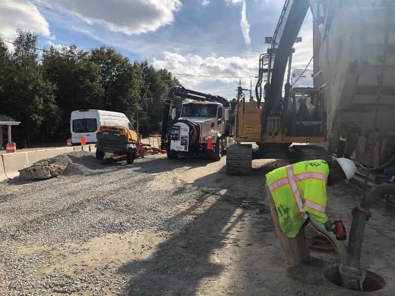 Roadwork scene with trucks and a worker in a neon vest near heavy machinery on a gravel road.