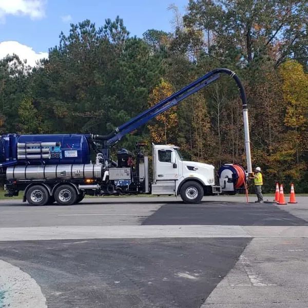 Blue vacuum truck and workers cleaning a roadside utility pole with cones nearby