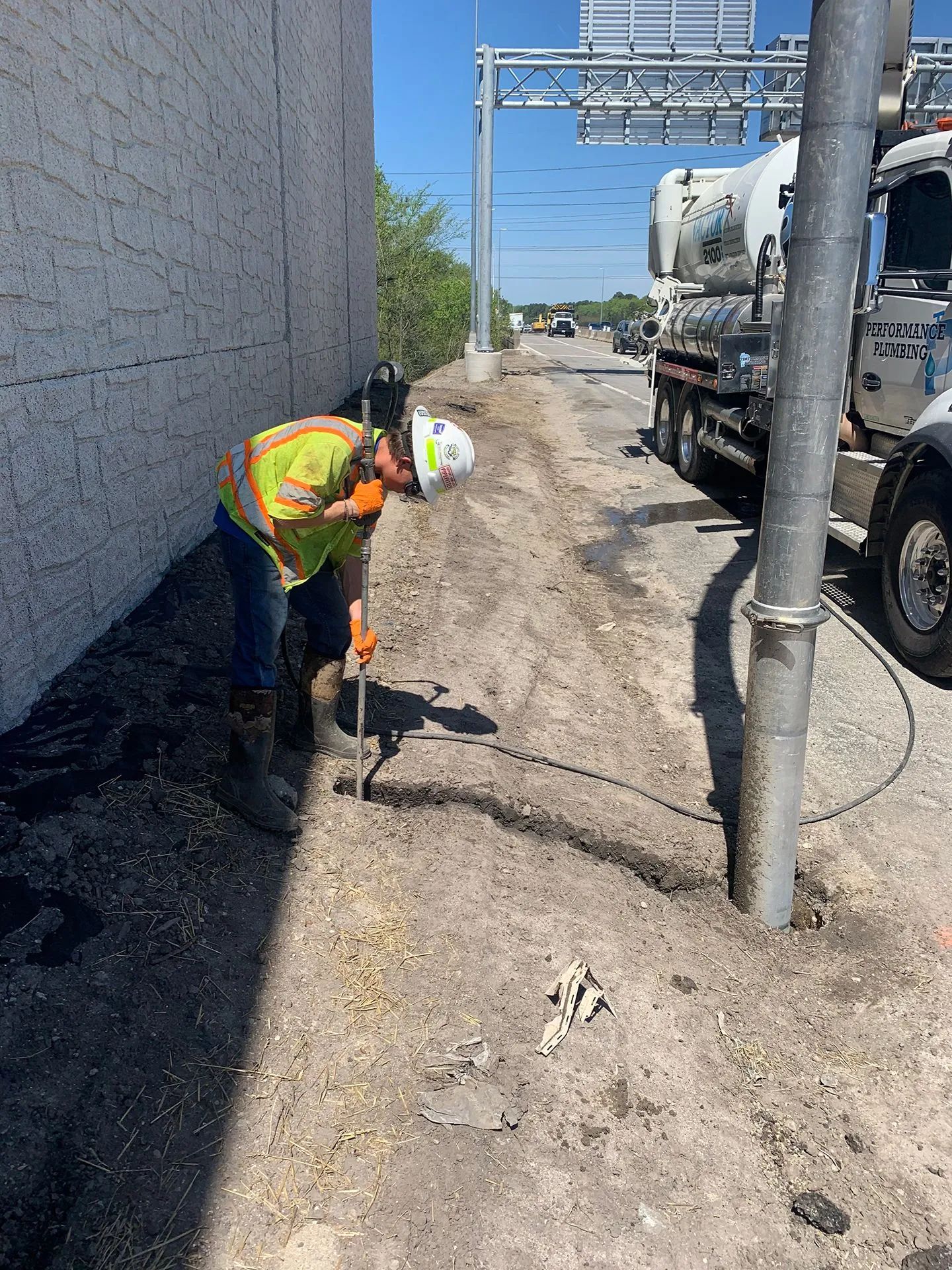 Worker in orange vest digging beside a building on a narrow service road with utility trucks and poles.