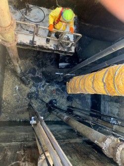 Worker in hard hat on a platform inside a deep excavation with pipes and muddy walls.