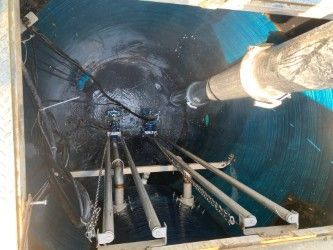 Inside a blue industrial tank, pipes and cables lead to a dark opening with a large pipe entering from above.