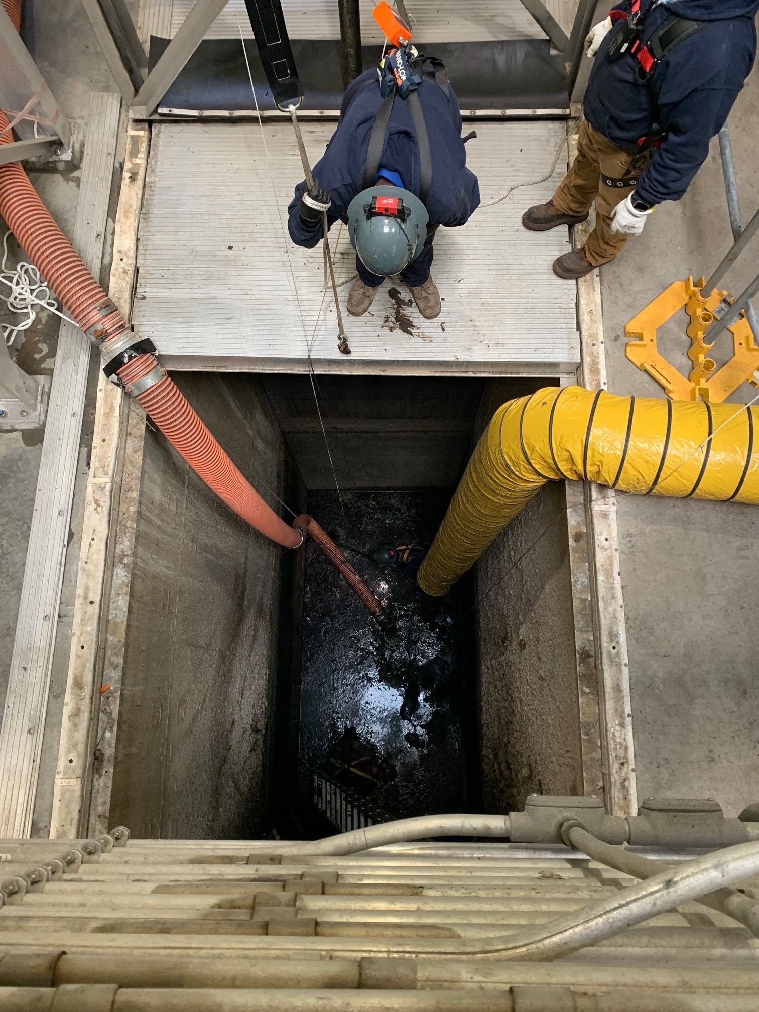 Workers descending a shaft via ladder and harness, with yellow ventilation ducting in an industrial site