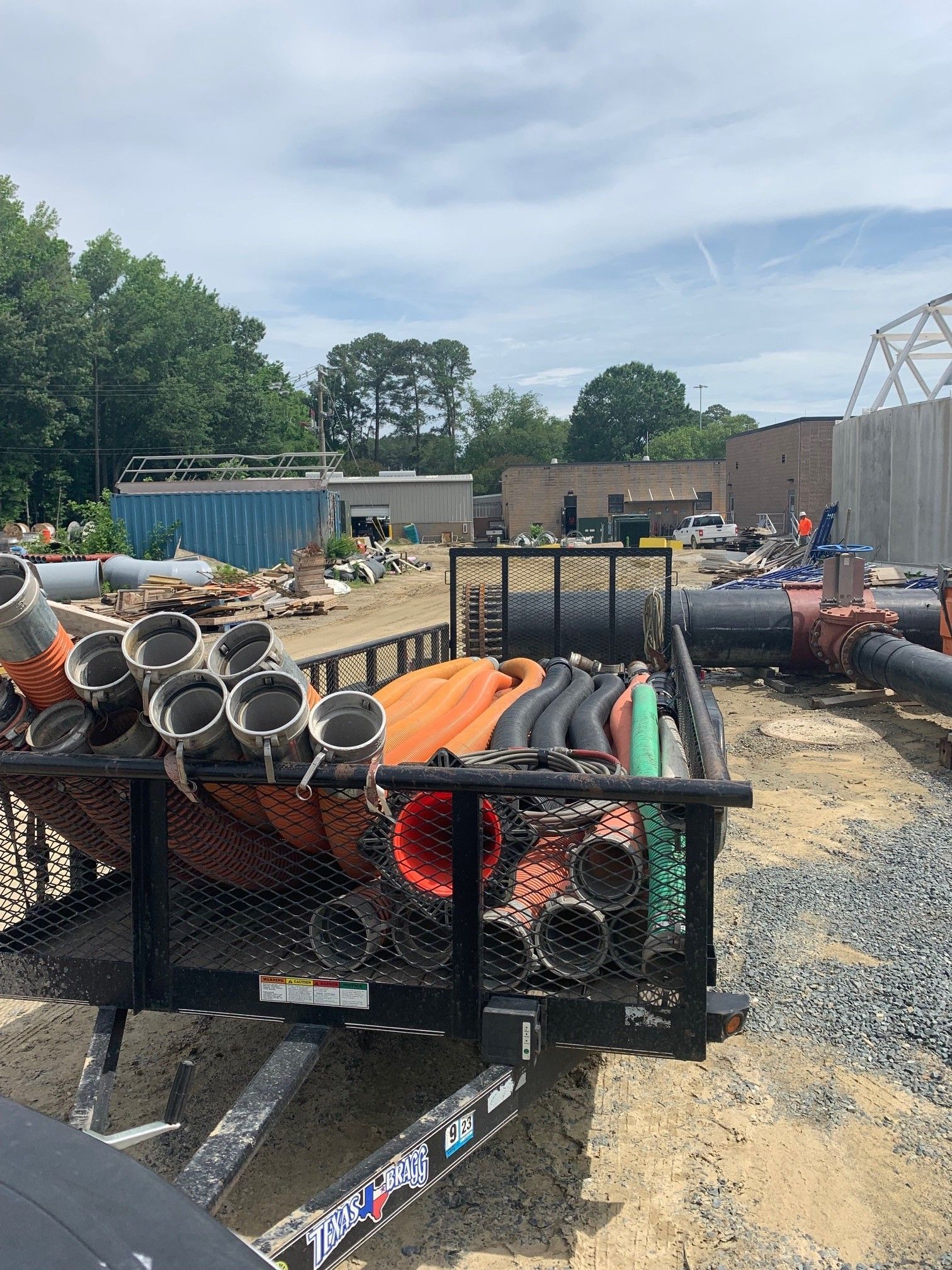 Construction site trailer loaded with pipes, orange safety mesh, and tools on a gravel lot