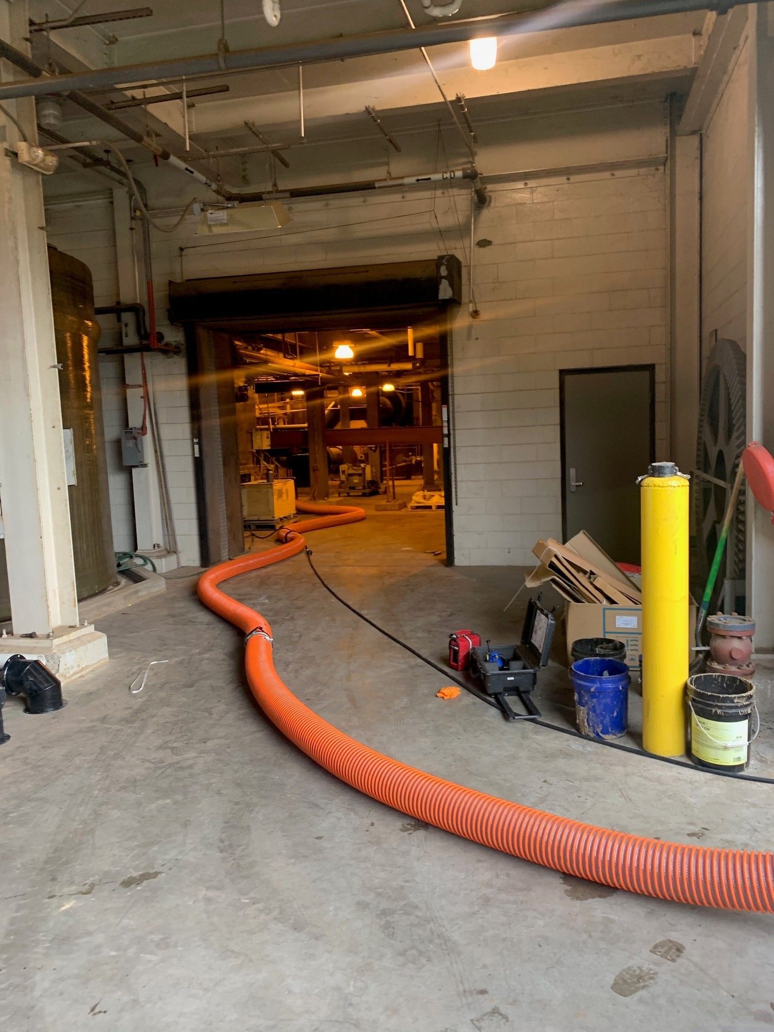 Industrial workspace with orange ventilation hose, tools, and supplies on concrete floor near open bay door