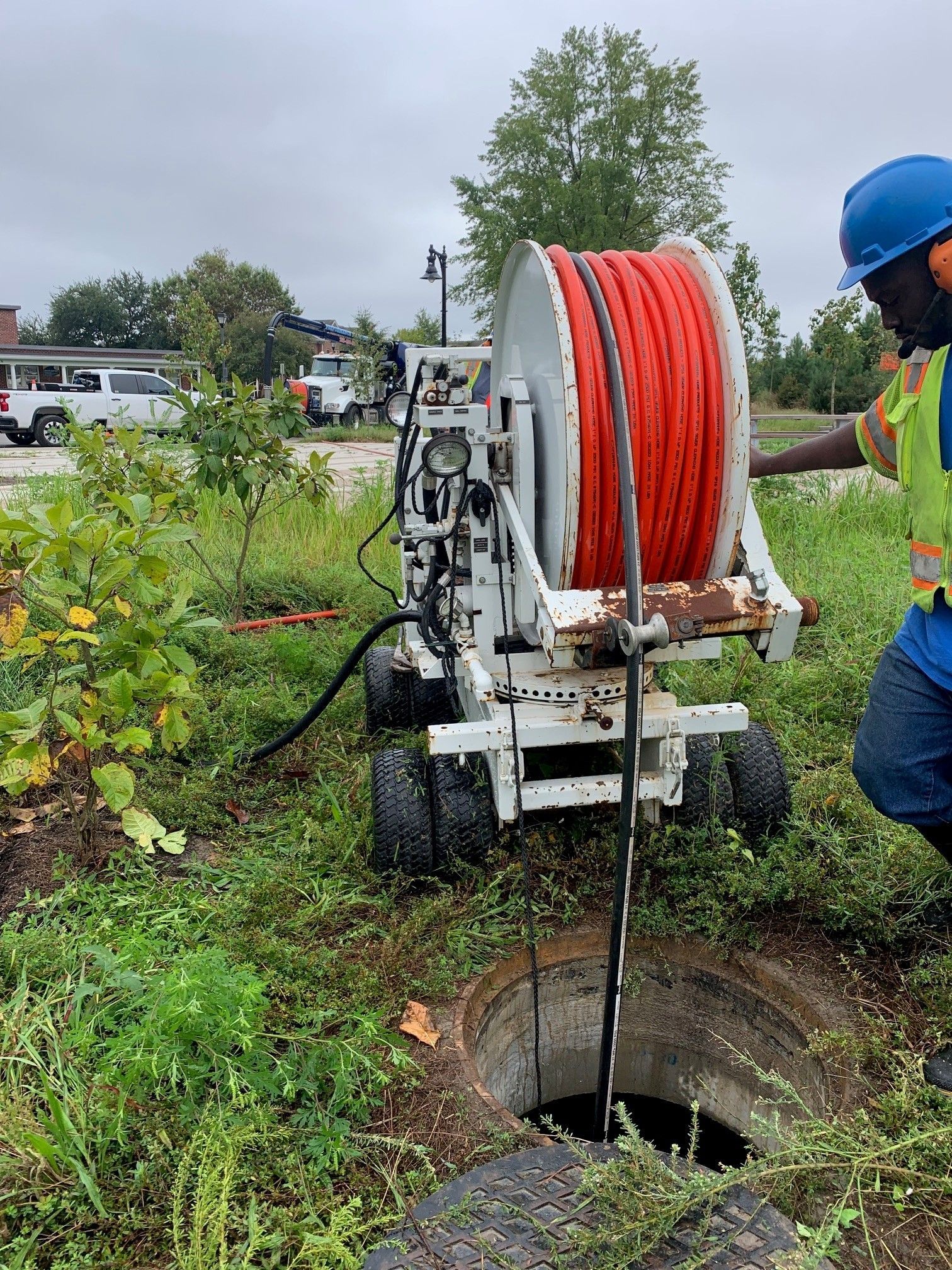 Worker in blue hard hat operating cable reel above an open manhole in a grassy roadside area