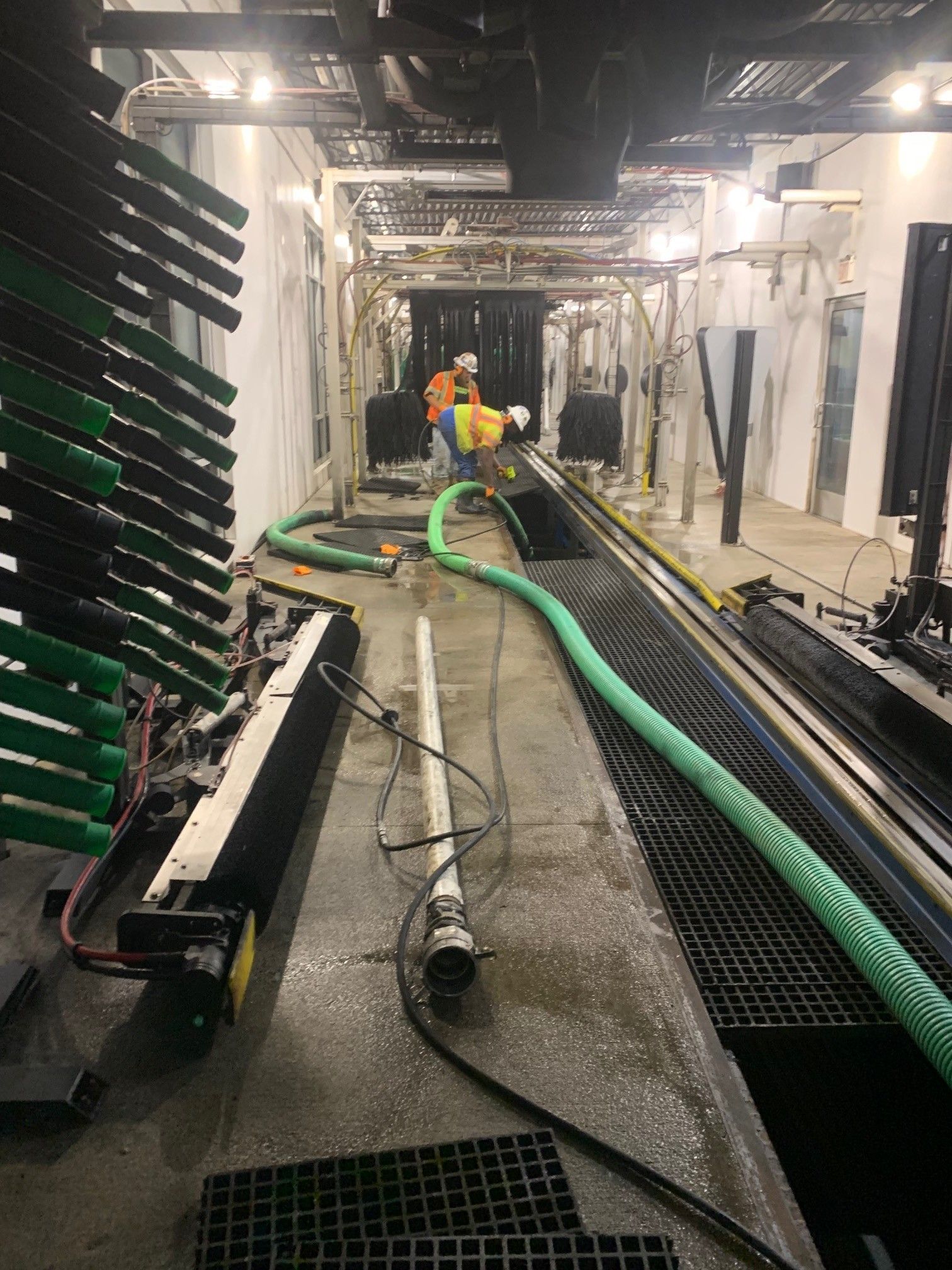 Workers in a lit industrial tunnel handle green hoses and cables on a wet floor