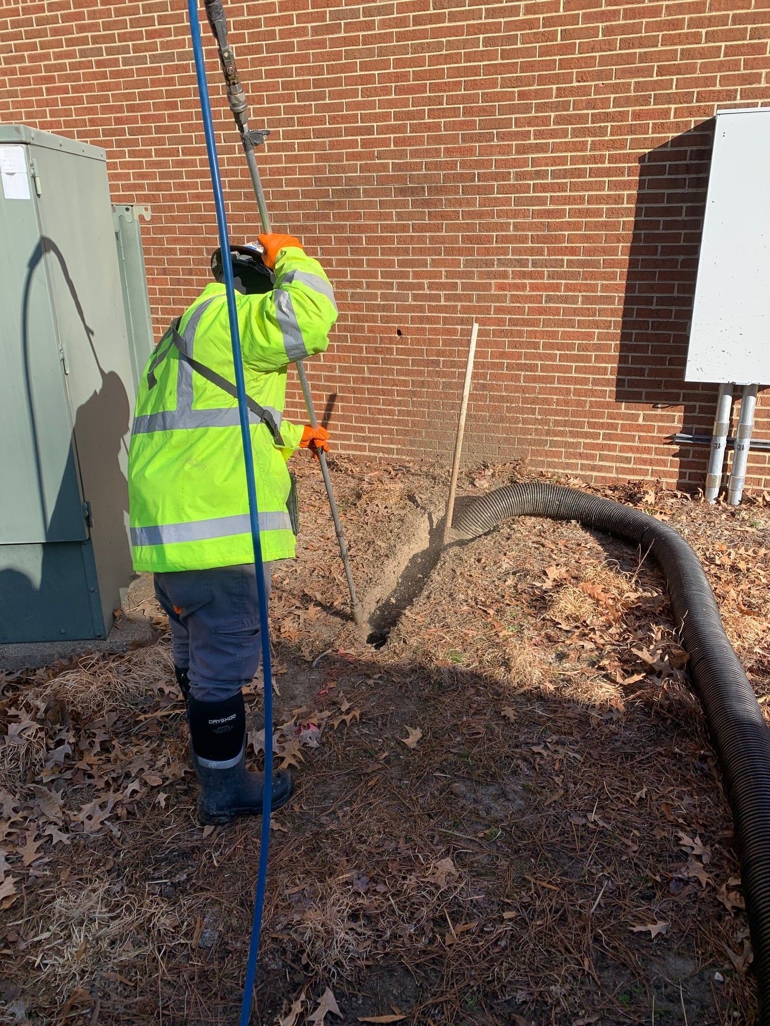 Worker in neon safety gear uses a tool beside a brick wall at a construction site.