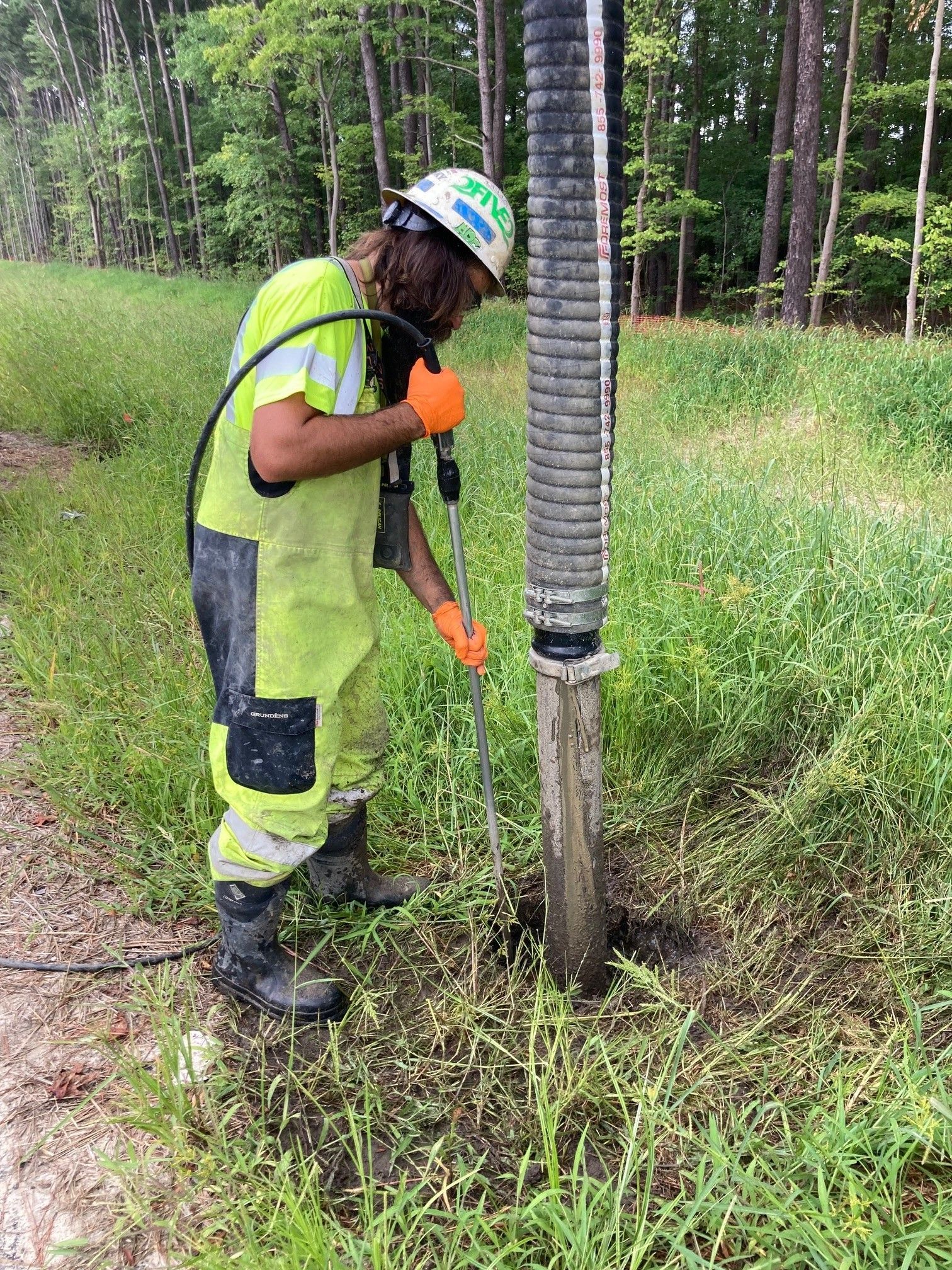 Worker in neon safety gear trimming grass around a utility pole in a wooded field.