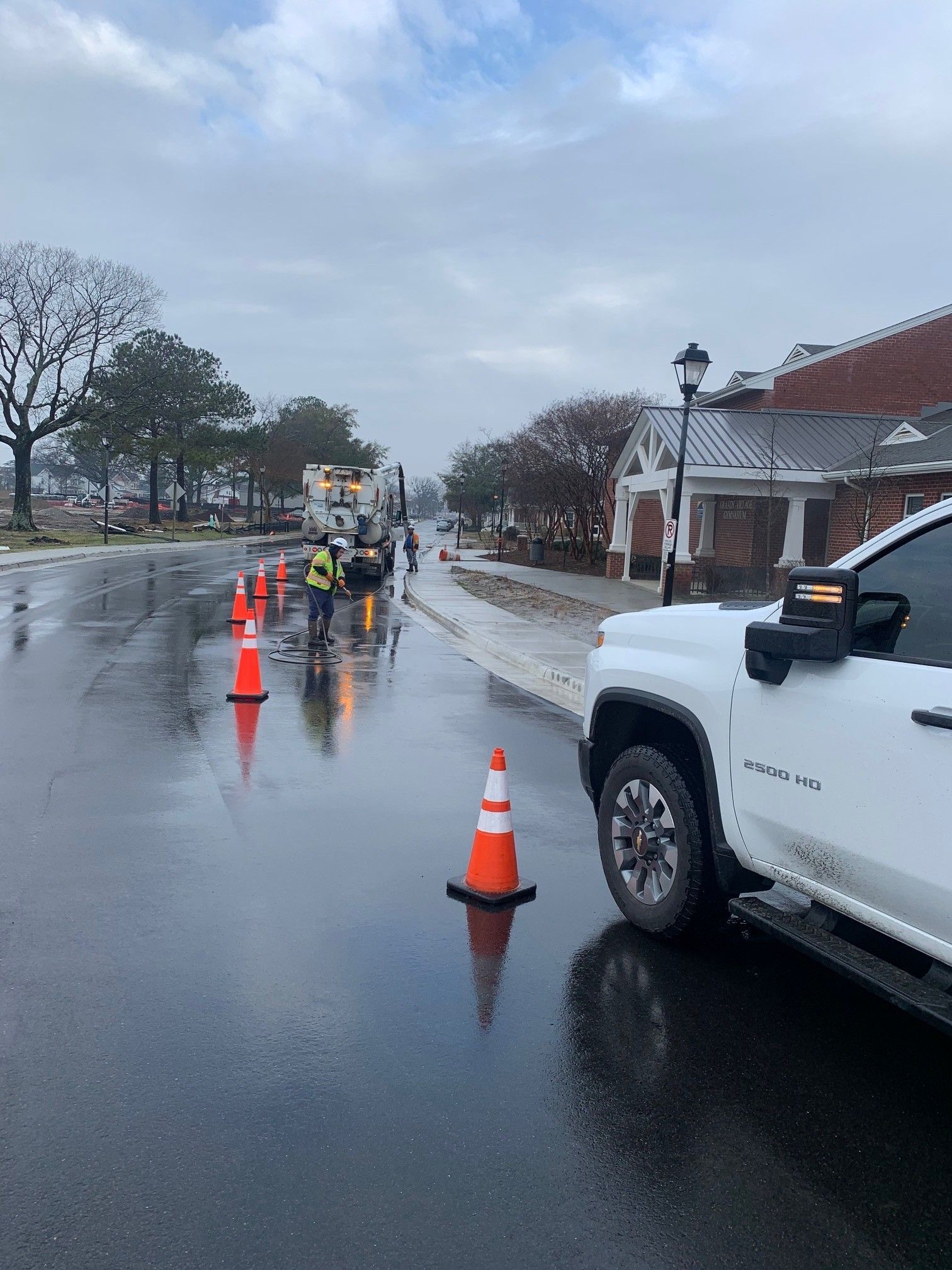 Wet street with orange cones and workers near a white truck beside a building on a rainy day