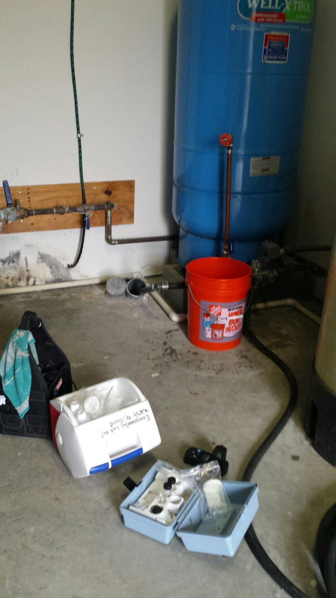 A red bucket is sitting on the floor next to a water tank.