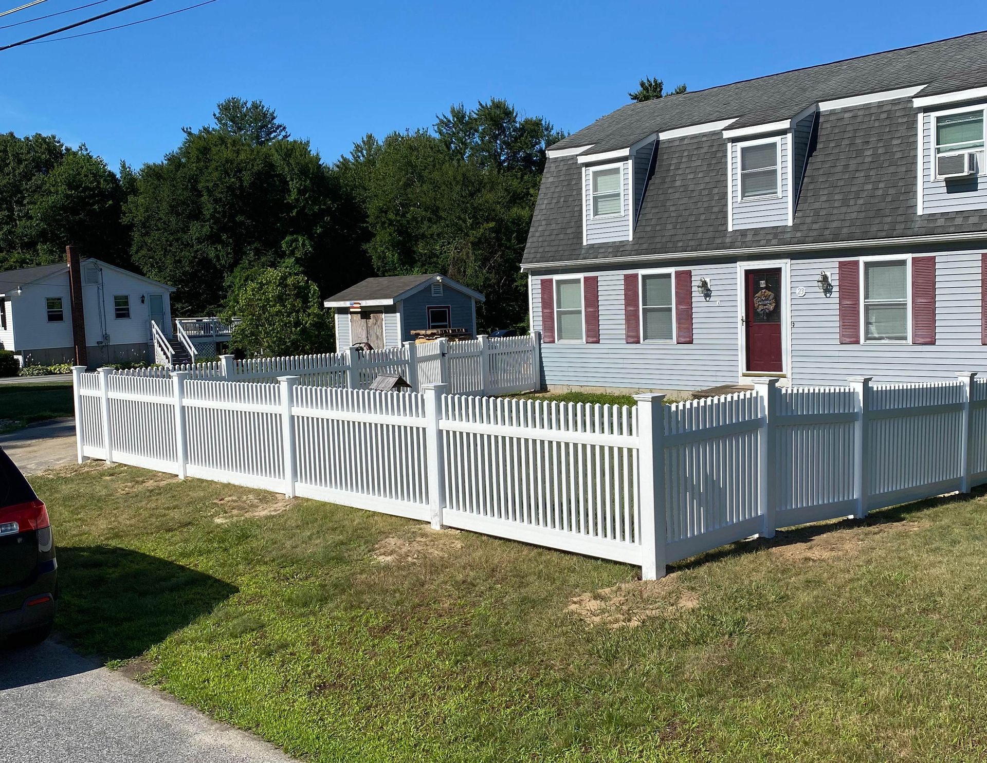 White picket fence bordering a lush green lawn, with trees and a house in the background.