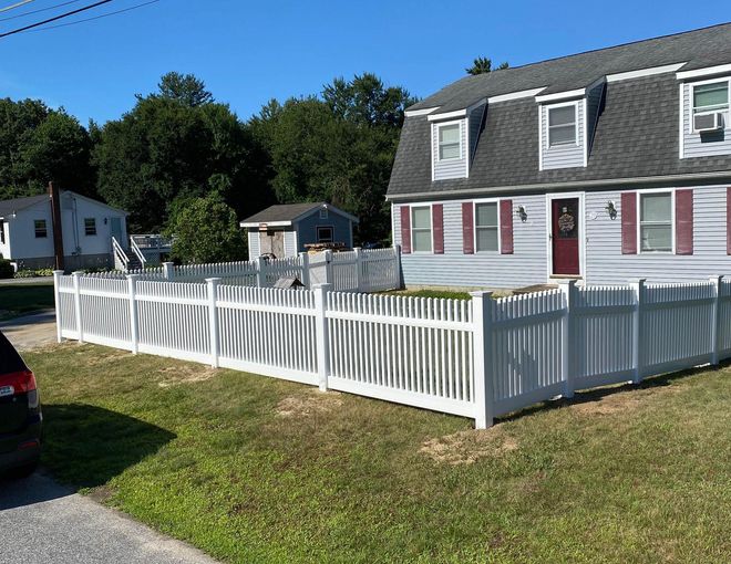 White picket fence bordering a lush green lawn, with trees and a house in the background.
