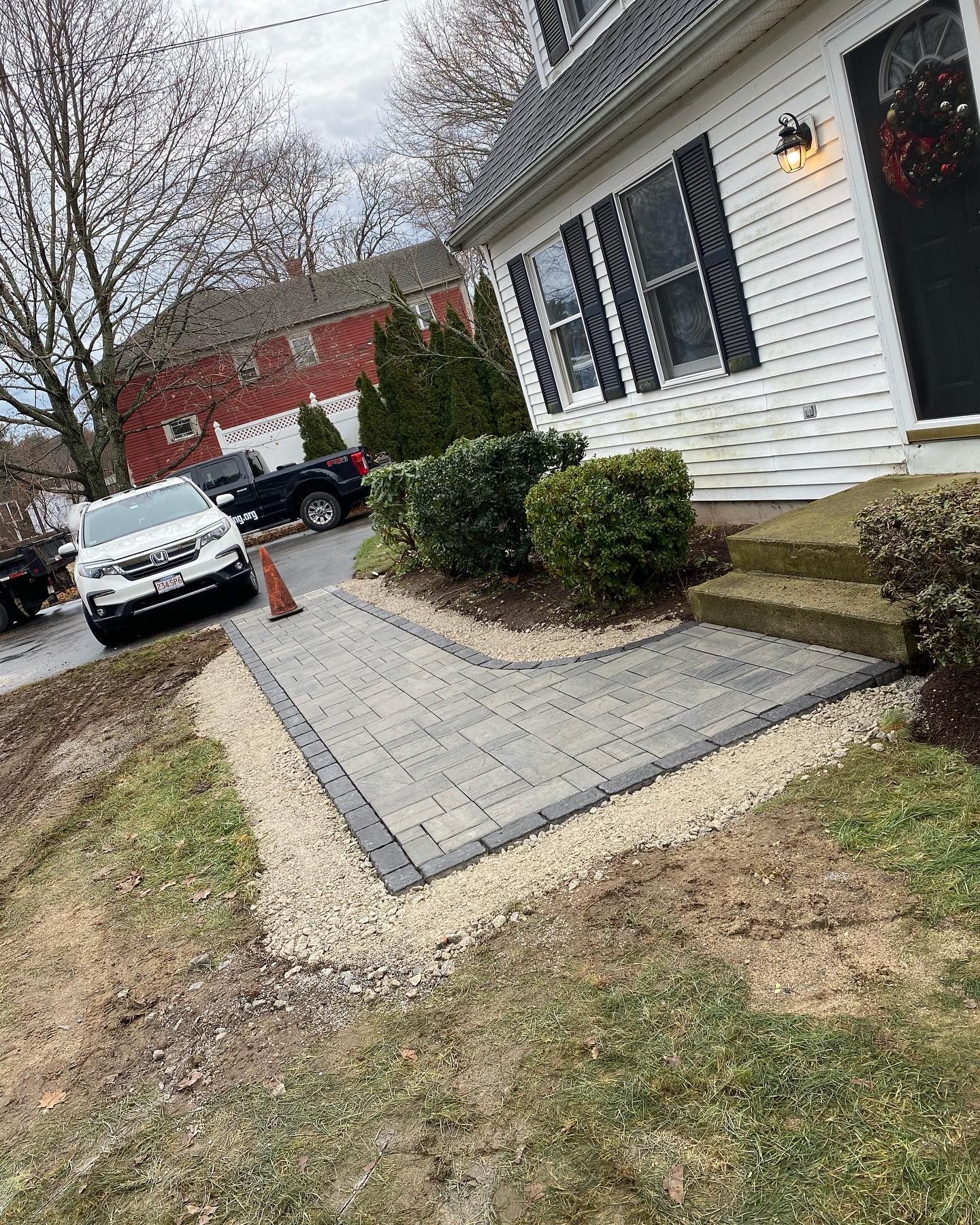 Brick walkway leading to a house with shrubs. Cars are parked in the background.