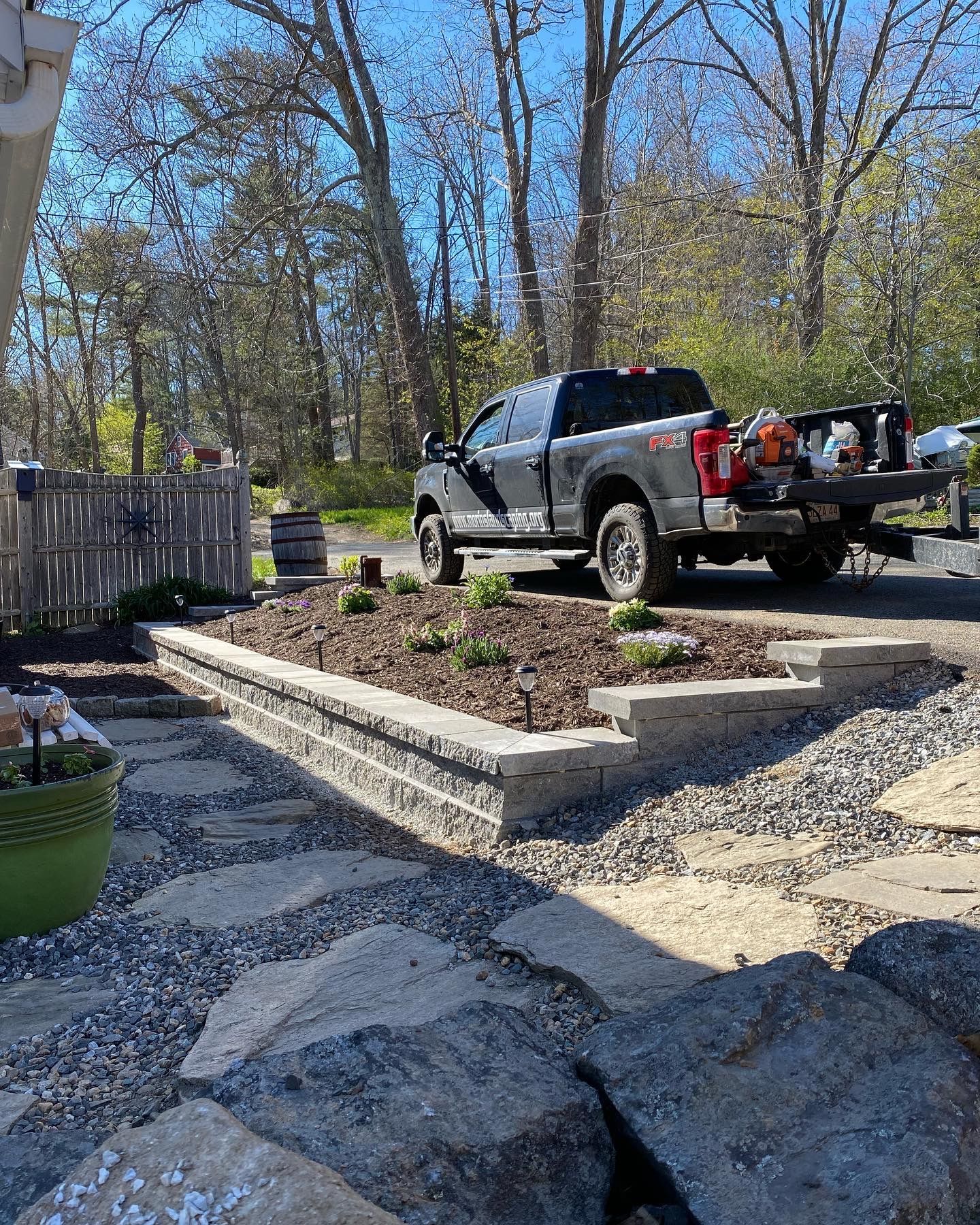 Grey truck parked near a raised garden bed with plants, stones, and a wooden fence on a sunny day.