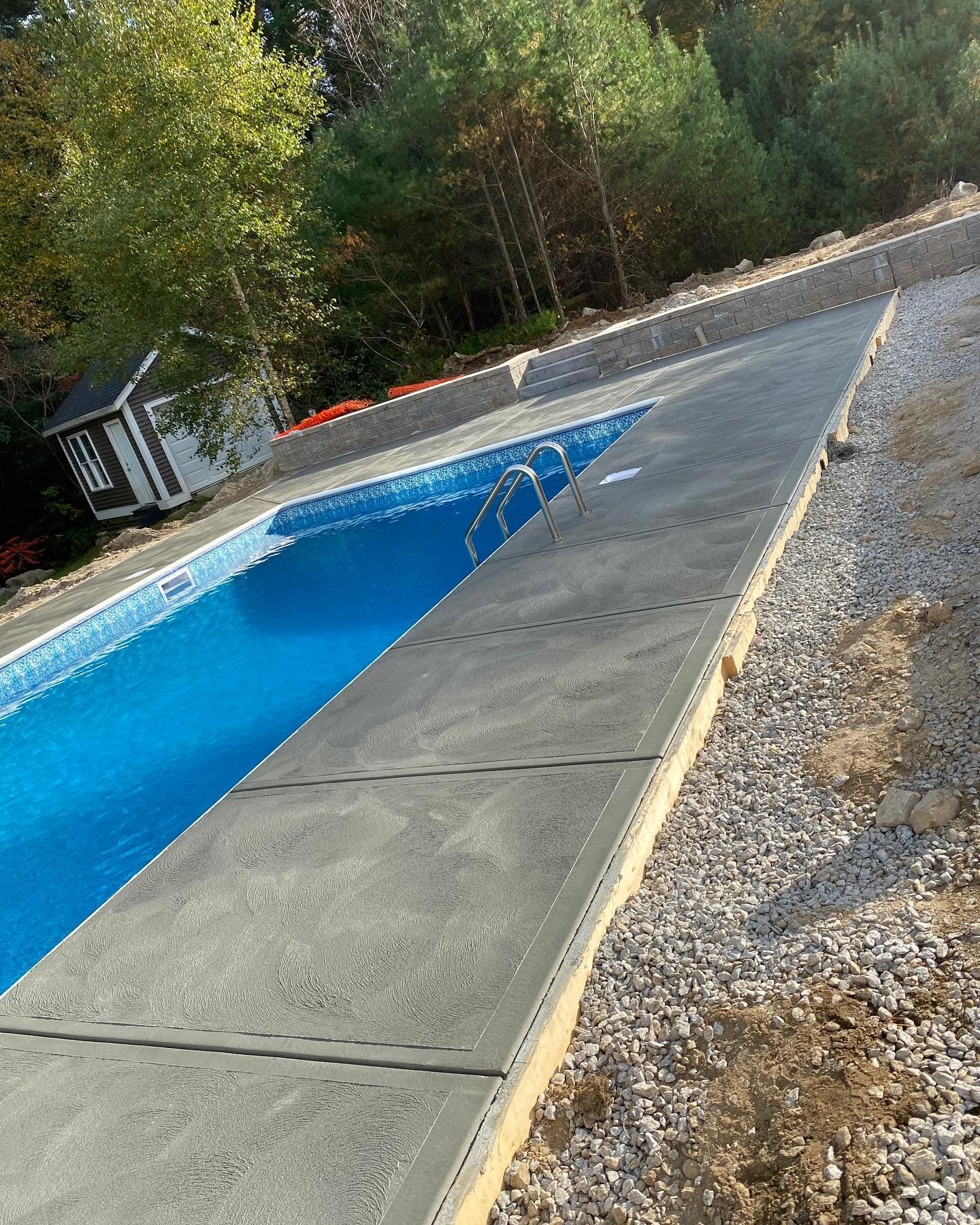Rectangular pool with blue water and gray concrete surrounding it. Small building, trees, and gravel also present.