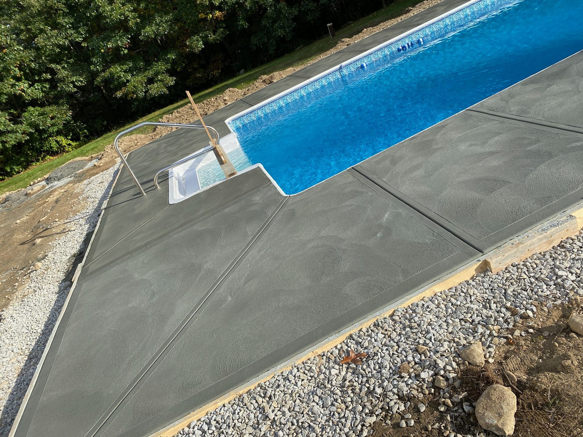 Concrete pool deck with blue water, surrounded by gravel and greenery.