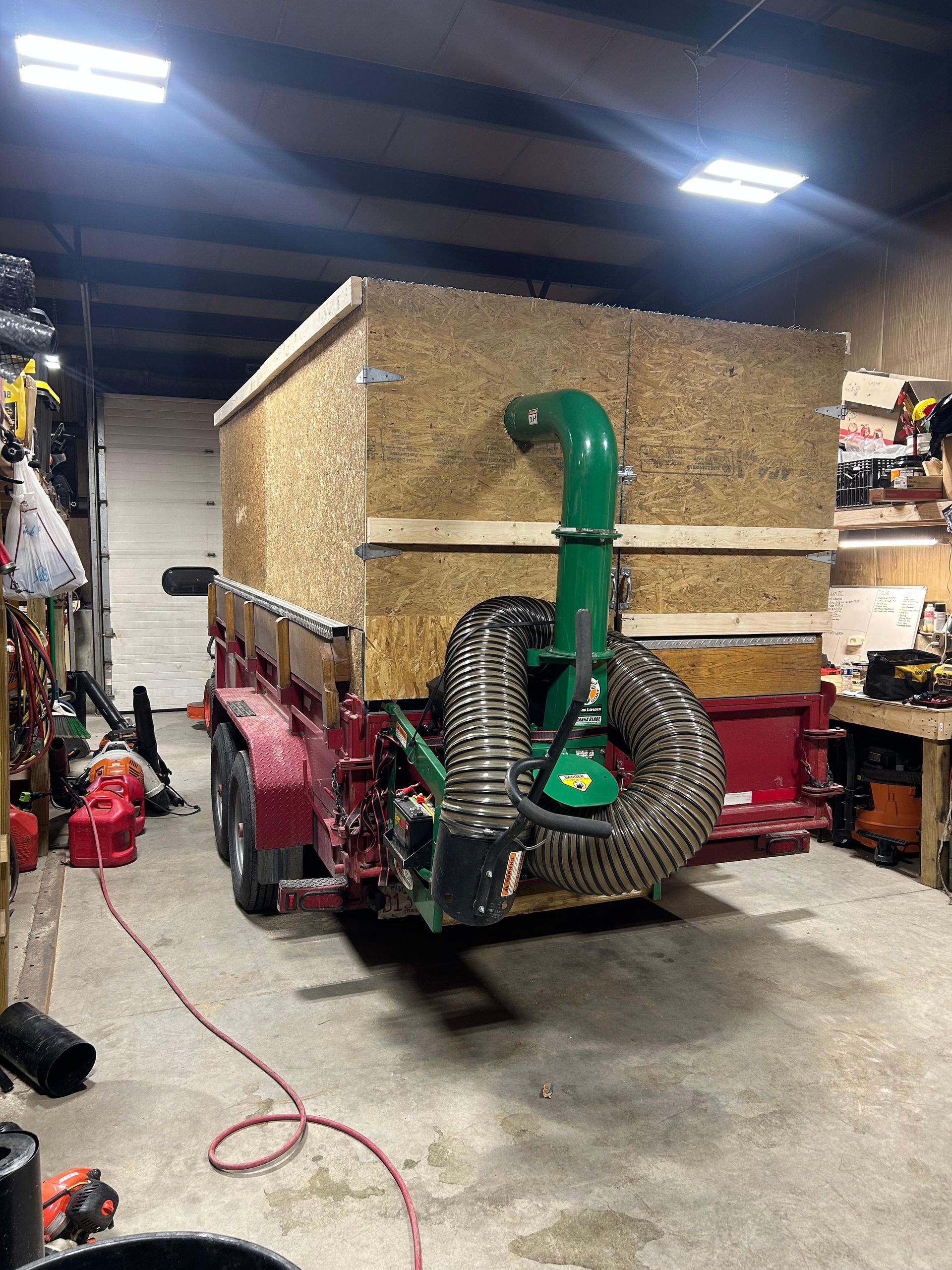 Leaf vacuum system attached to a trailer inside a workshop. Red trailer, tan wood box, green hose.