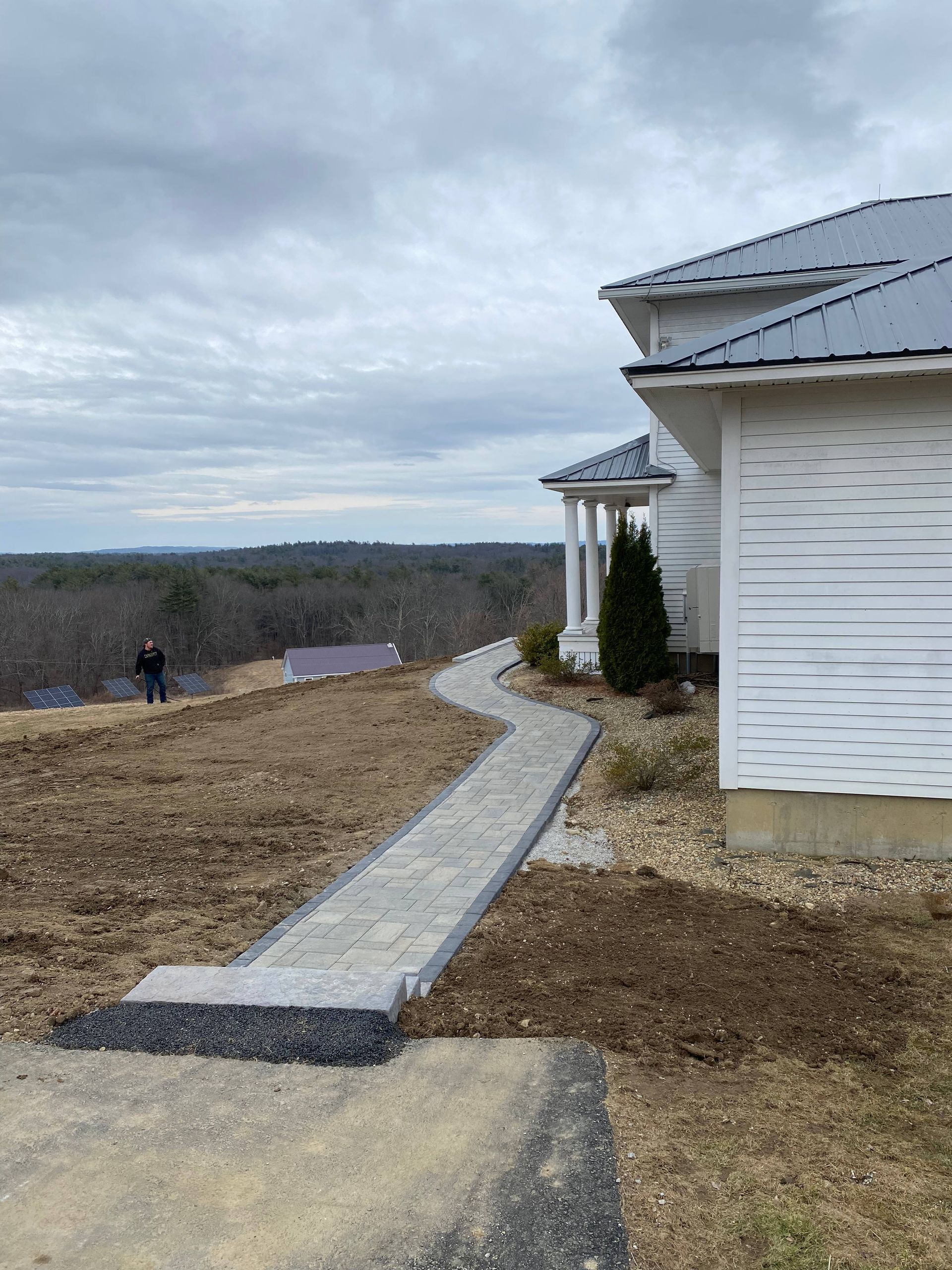 Stone walkway leading to a white house with a gray roof, overlooking a wooded landscape under a cloudy sky.