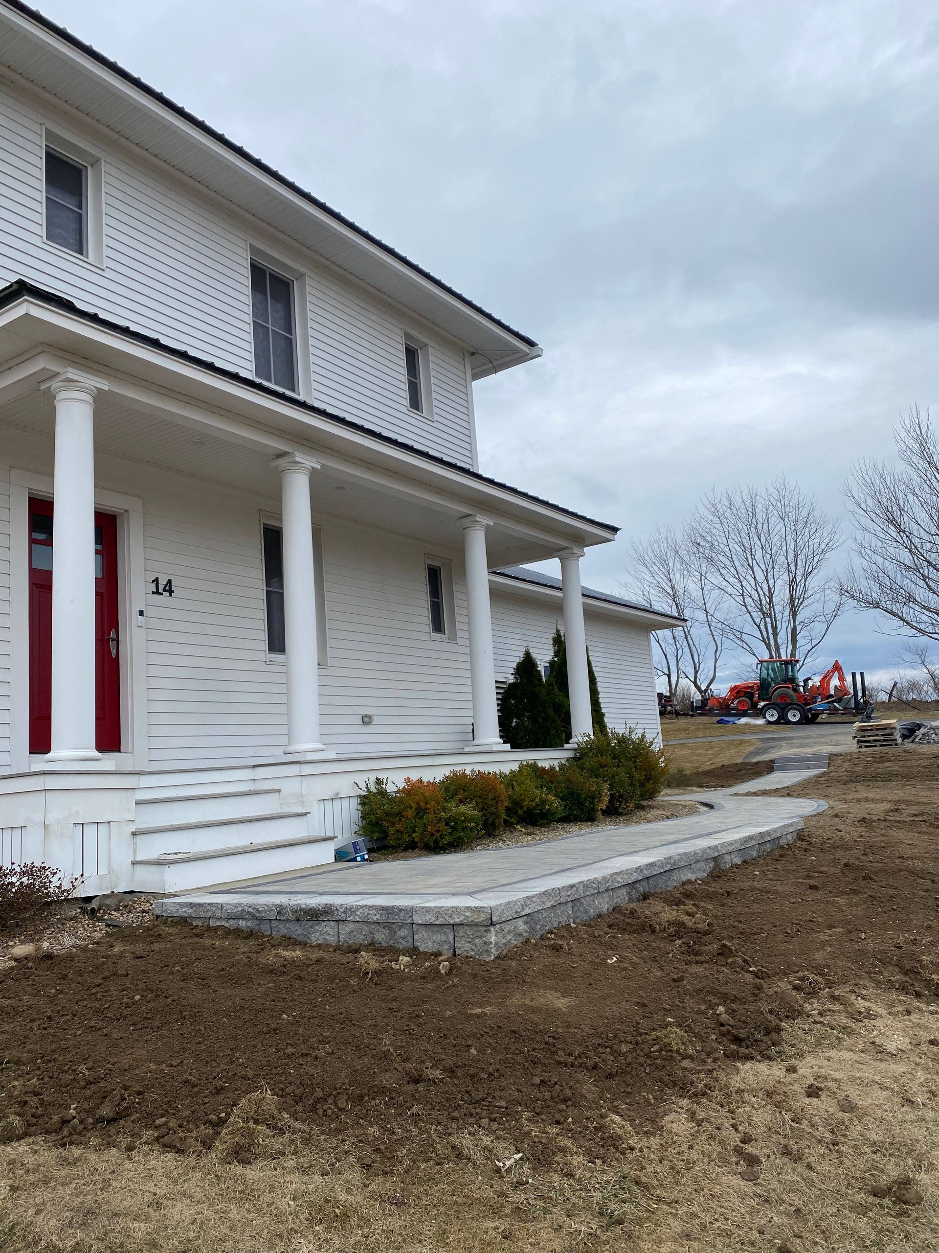 White two-story house with porch. Red door, gray stone border, and bare dirt in front. Overcast sky.
