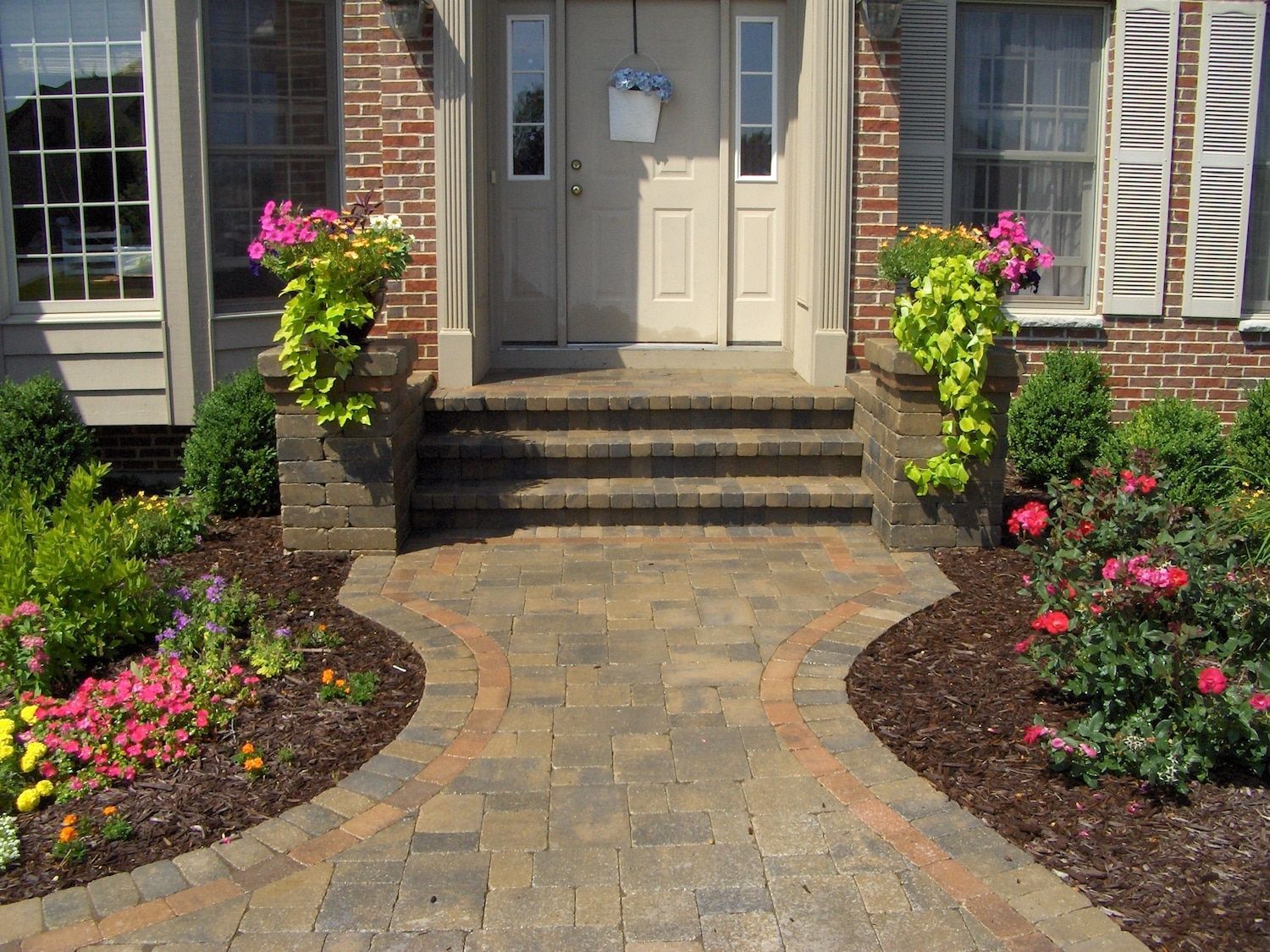 Brick walkway leading to a house with steps and planters, lined with flowers.
