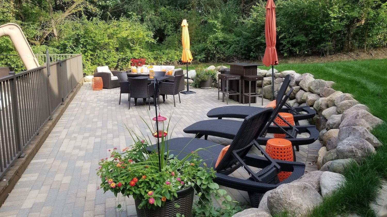 Outdoor patio with lounge chairs, dining set, bar, and rock border; lush green lawn and trees.