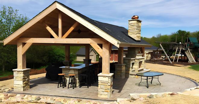 A timber-framed outdoor pavilion with a stone fireplace, bar seating, and a paved patio area in a backyard.