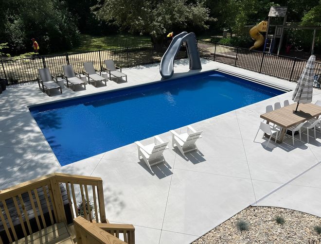 An aerial view of a bright blue rectangular swimming pool with a slide, lounge chairs, and an outdoor dining set on a patio.