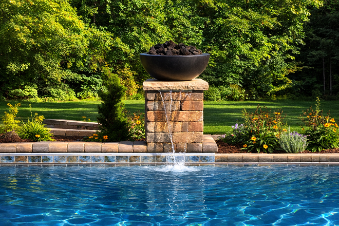A brick fountain with a black basin and fire rocks sits on the edge of a bright blue swimming pool by a lush, green forest.