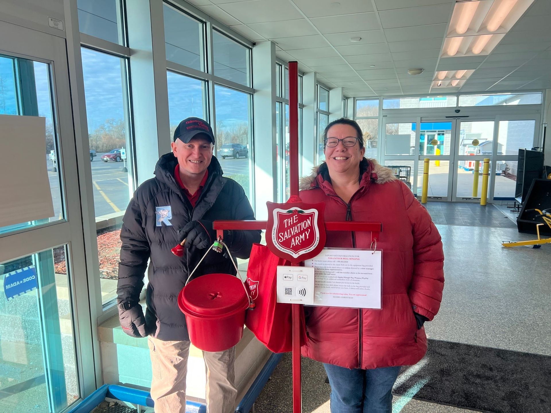 Two people stand by a Salvation Army bell-ringing station. A red bucket and sign are visible.