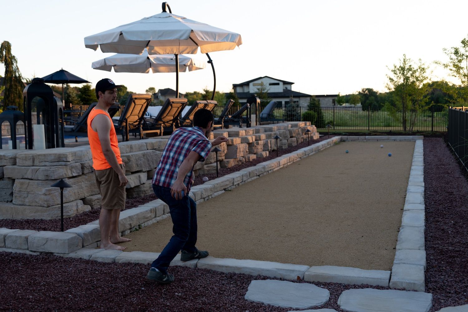 Two men playing bocce ball on a gravel court at dusk; one throws a ball, the other watches.