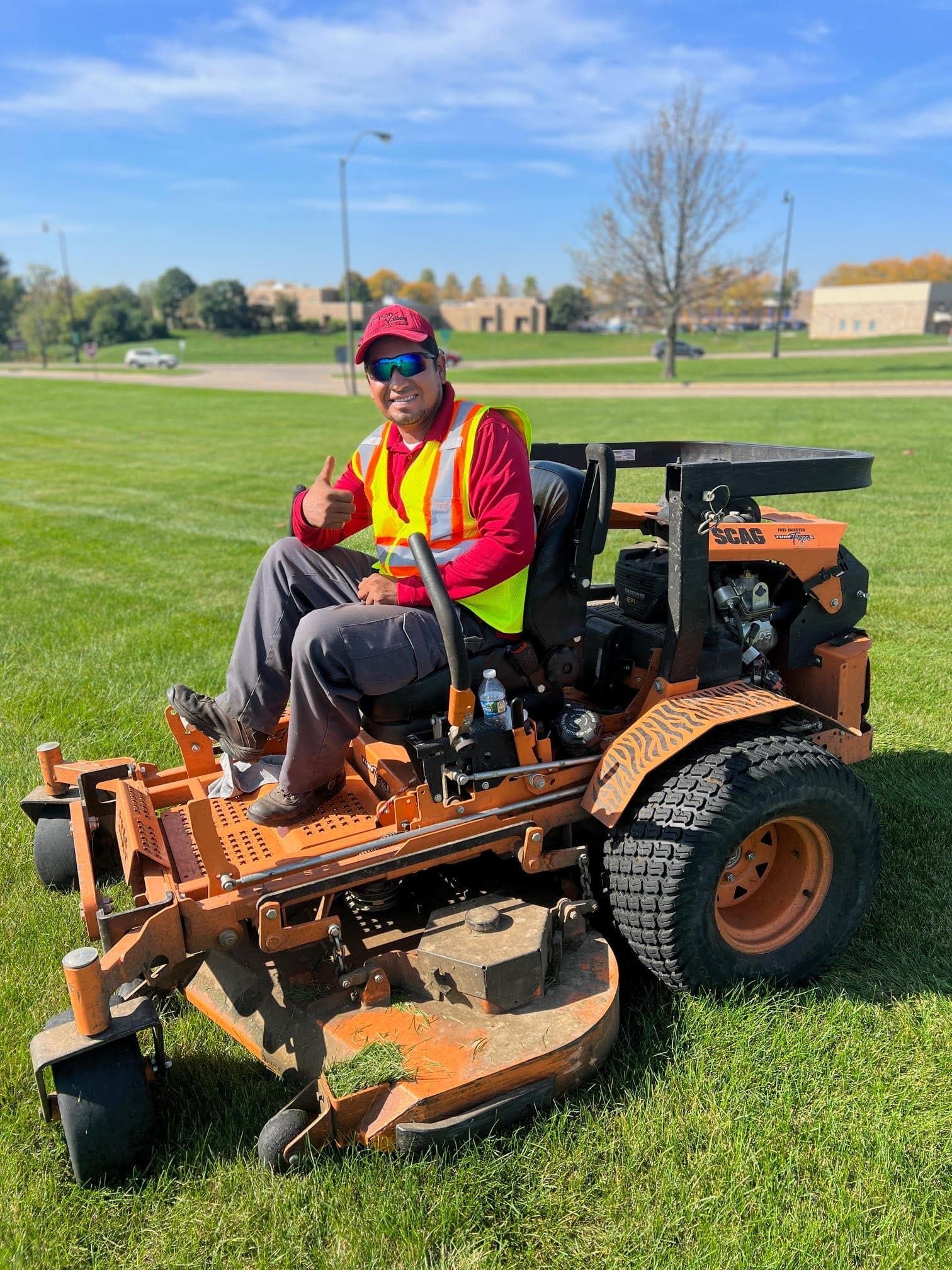 Man on orange lawnmower, giving thumbs up on a green field under a blue sky.
