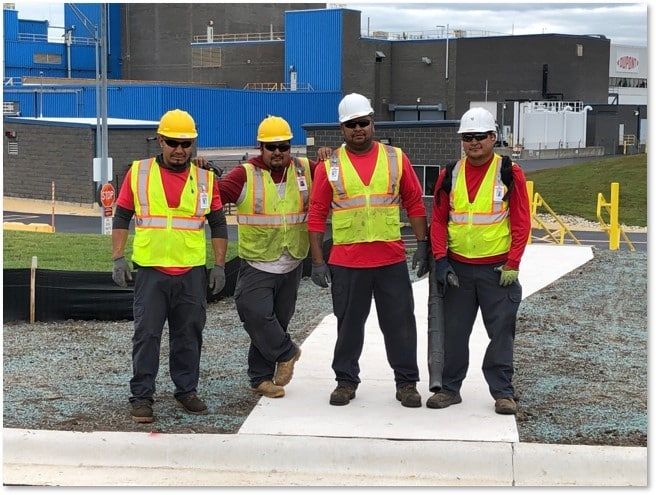 Four construction workers in safety vests and hard hats stand outside a building.