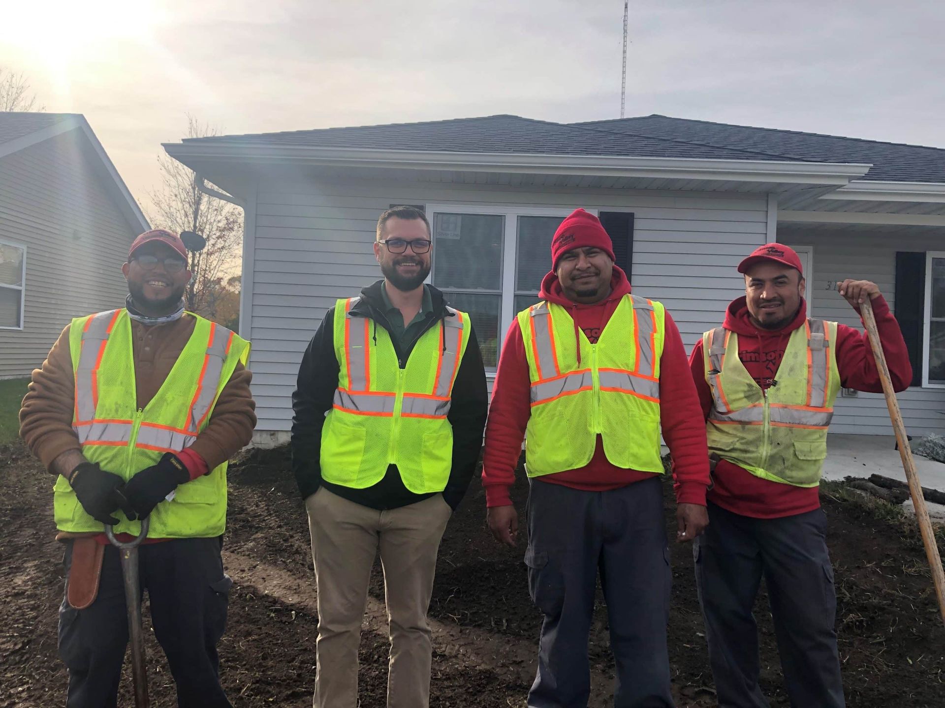 Four construction workers in safety vests stand in front of a house, one holding a shovel.