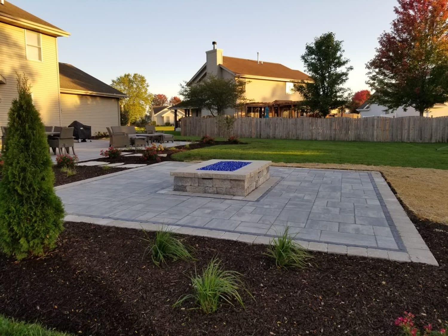 Stone patio with fire pit, surrounded by mulch, plants, and grass lawn; houses in background.