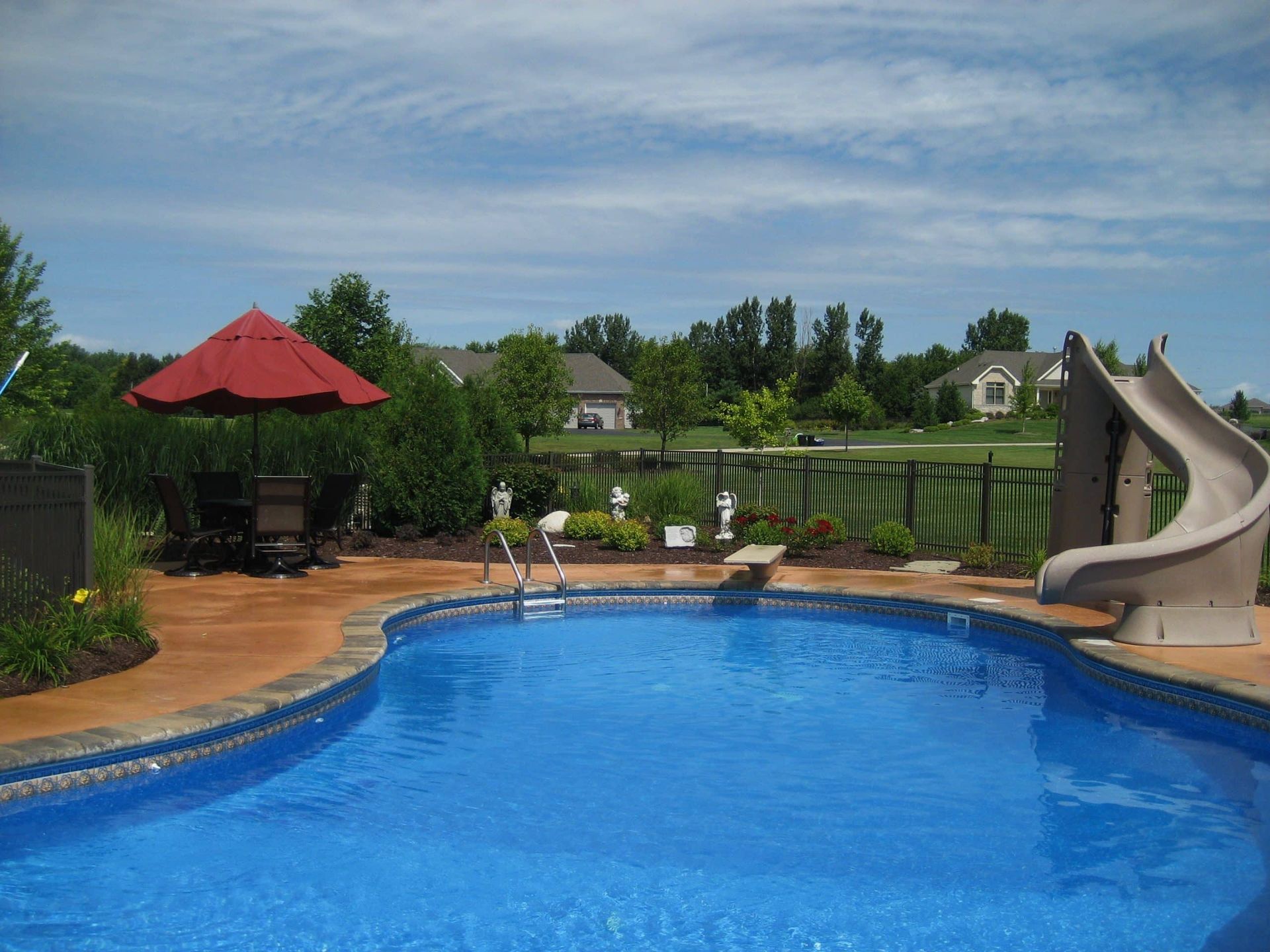 A swimming pool with a slide, umbrella, and seating area under a sunny, blue sky.