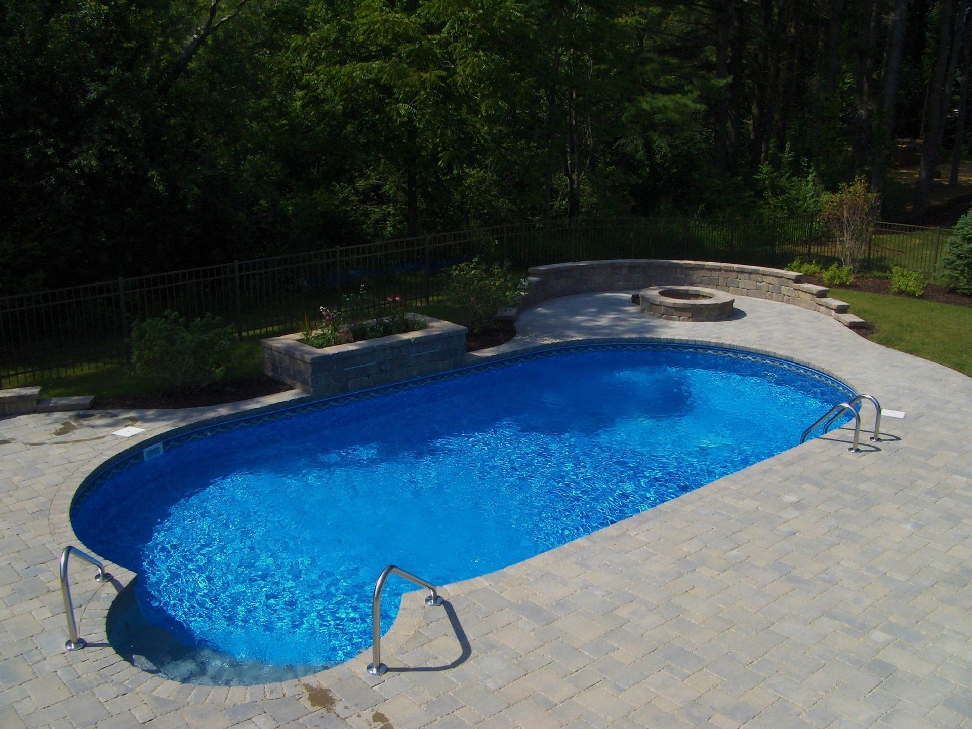 Oval swimming pool with blue water surrounded by stone patio and landscaping.