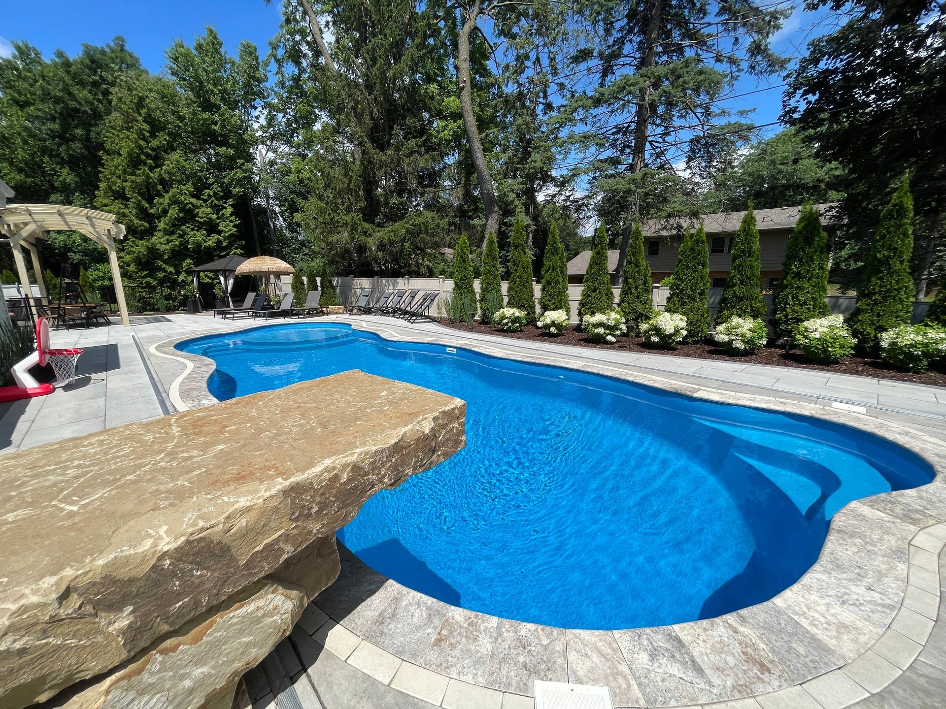 Swimming pool with blue water and stone diving board in a backyard. Trees and greenery surround the pool.