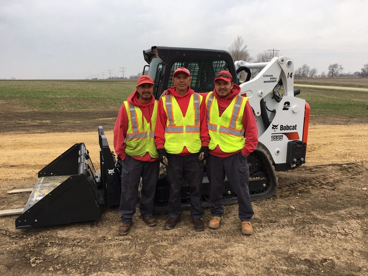 Three workers in red and yellow vests stand next to a Bobcat skid steer on a dirt path.