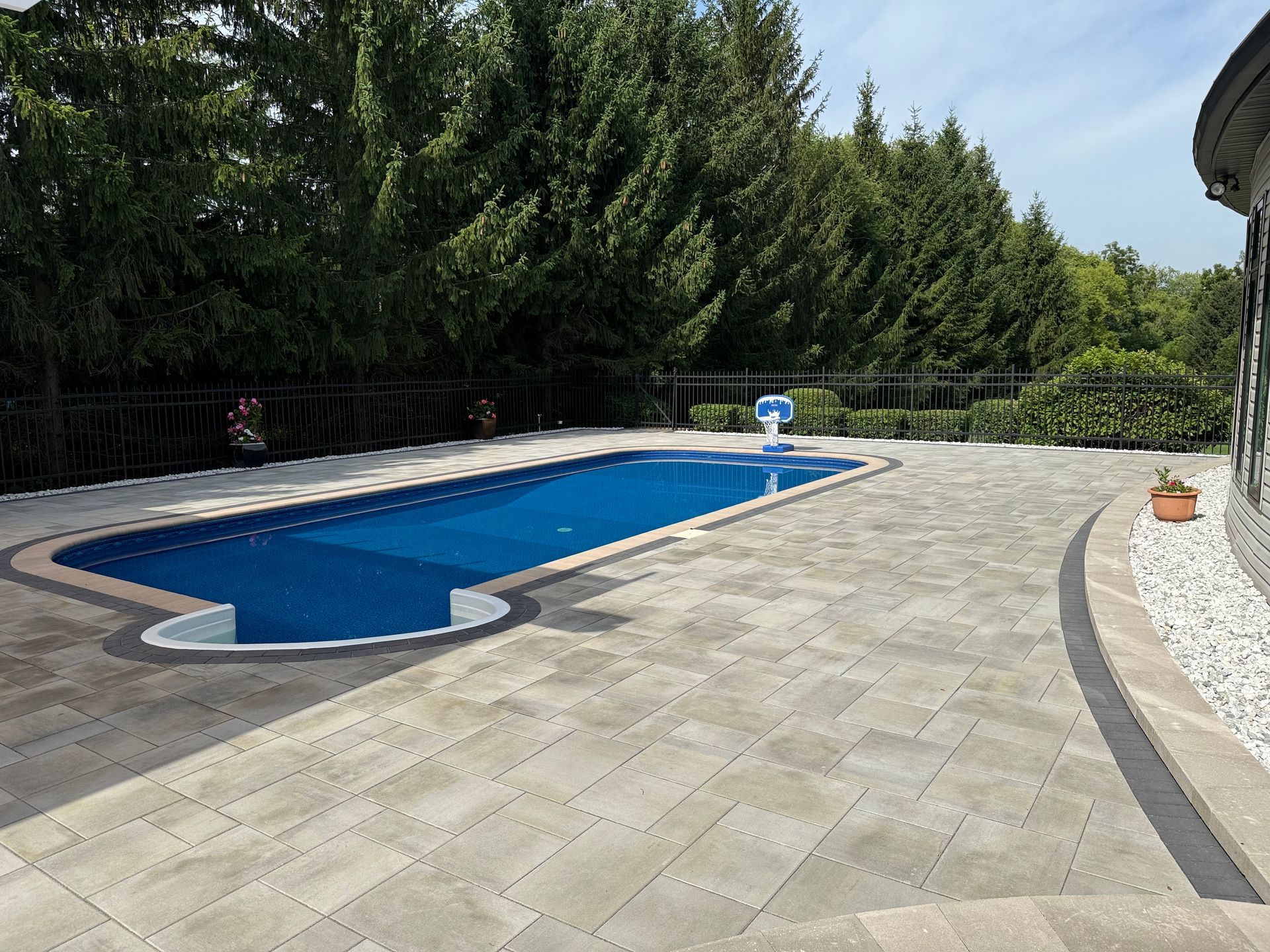 Backyard with rectangular pool covered by blue tarp, surrounded by paved patio. Trees in background.