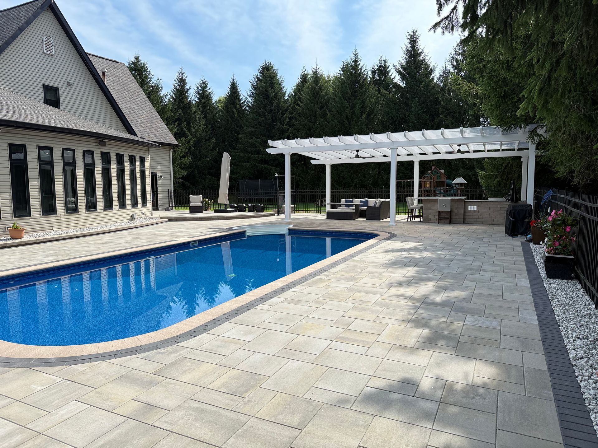 Backyard with a pool, pergola, and house. Blue pool, beige patio, and green trees are visible.