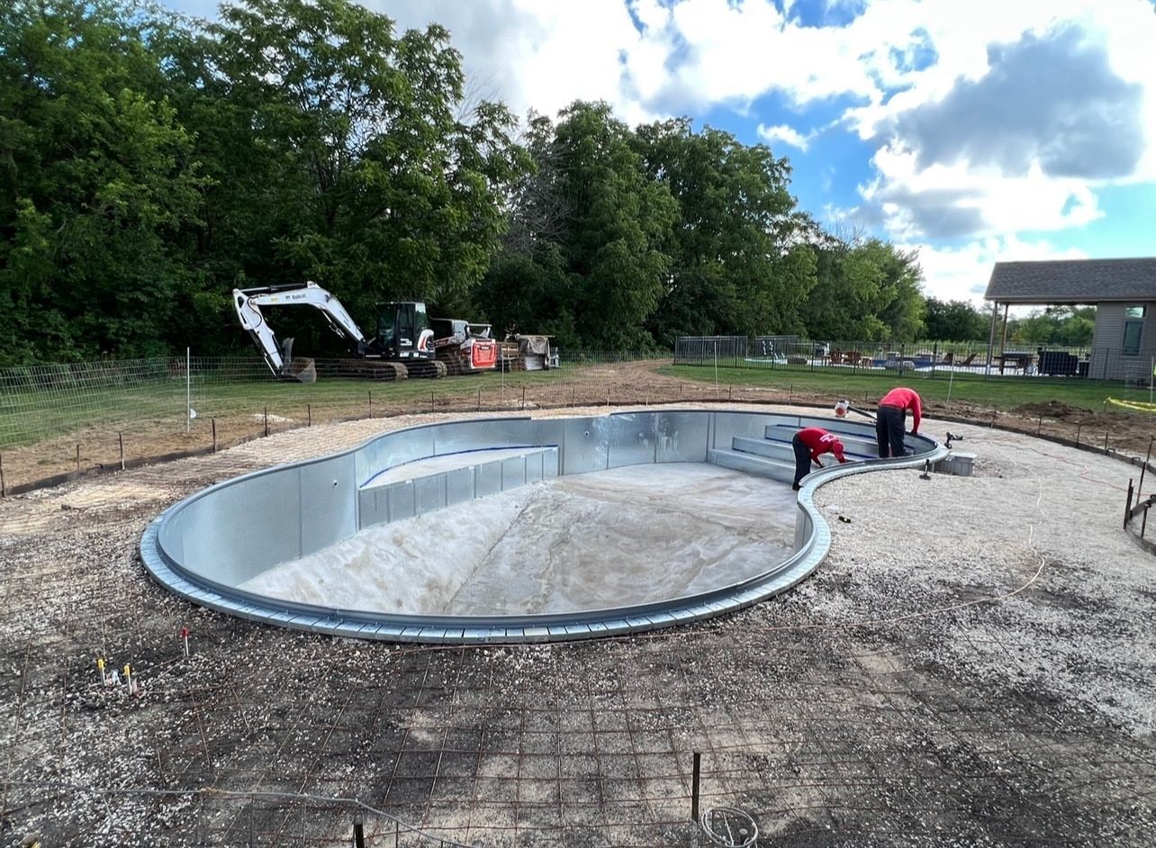 Pool under construction in a yard; workers, equipment, and a blue sky are visible.