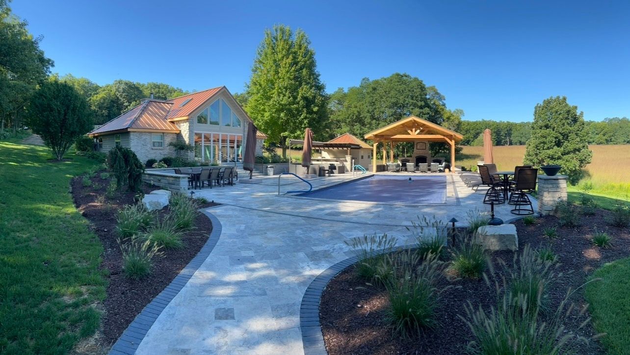 Backyard with pool, stone patio, outdoor kitchen, and house on a sunny day.