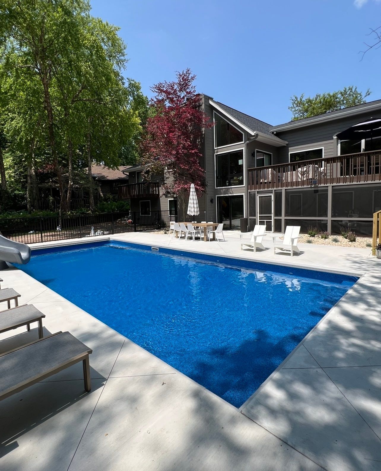 Blue swimming pool in front of a gray house with a deck; sunny day.