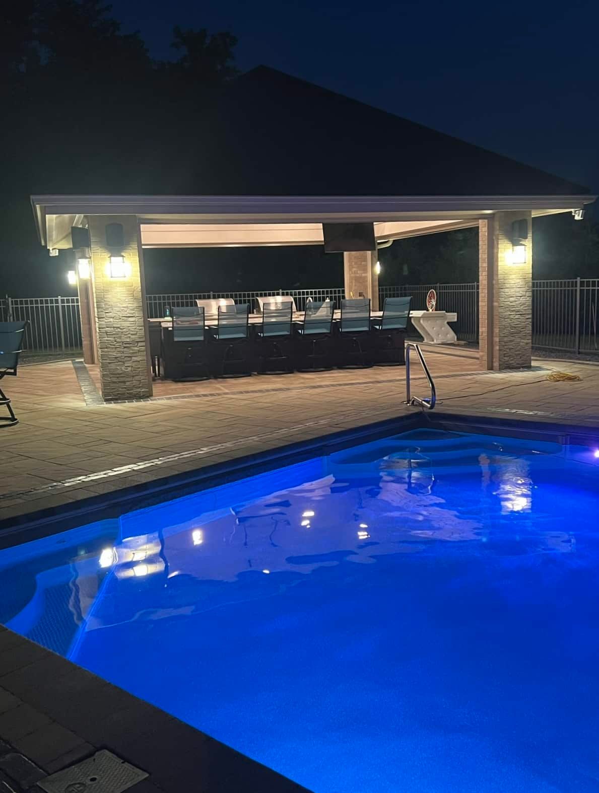 Poolside bar area lit at night, blue pool water, stone columns, chairs, lights.