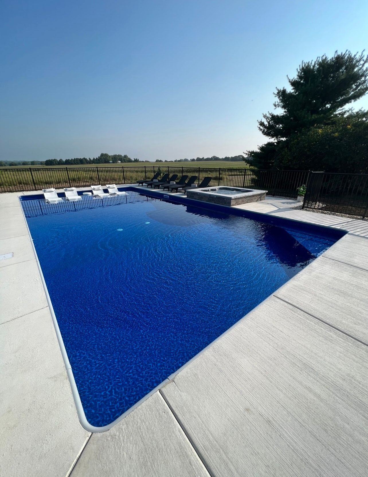 Blue swimming pool with light concrete border, steps, and distant vineyard under a blue sky.
