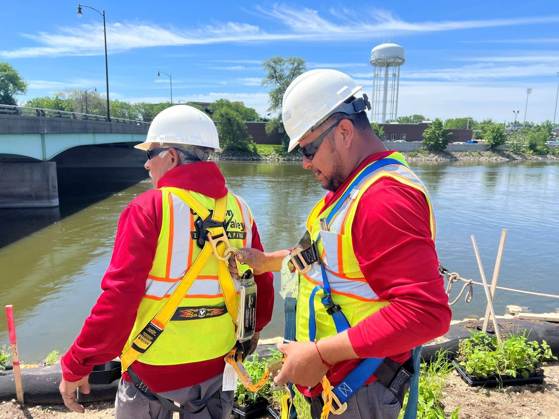Two workers in safety gear by a river. One adjusts the other's harness. A bridge and water tower are in the background.