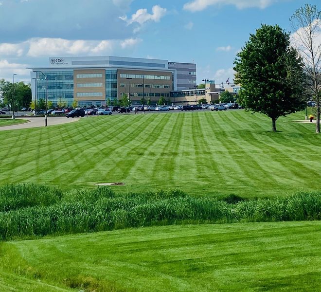 A large, neatly mowed lawn with prominent striped patterns leads to a modern multi-story office building under a blue sky.