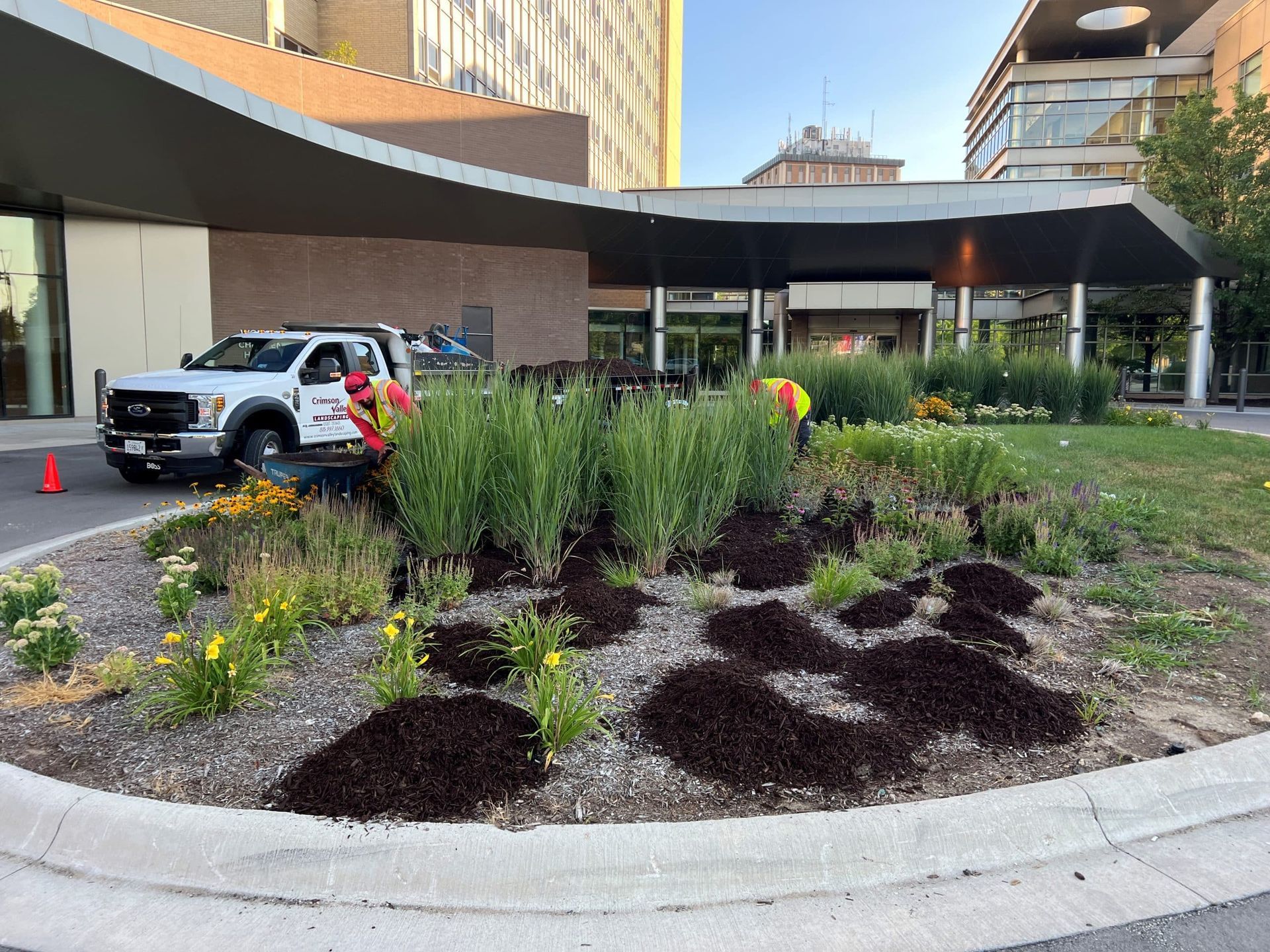 Workers tending a garden bed at a building entrance; a white truck with a company logo is parked nearby.