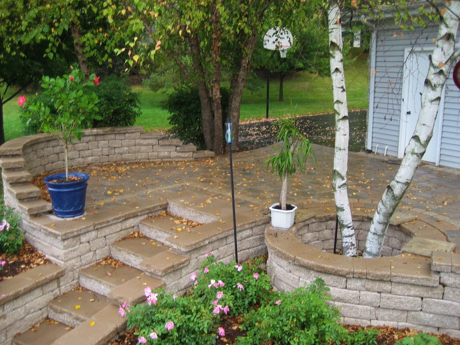 Stone patio with tiered levels, trees, and potted plants in a backyard.