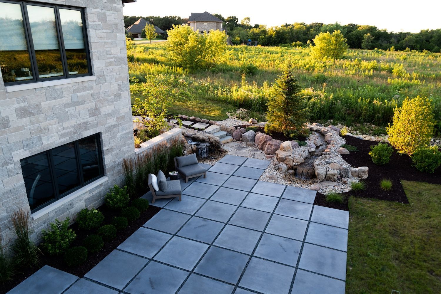 Patio with grey pavers, two chairs, and a small rock garden near a modern home's exterior.